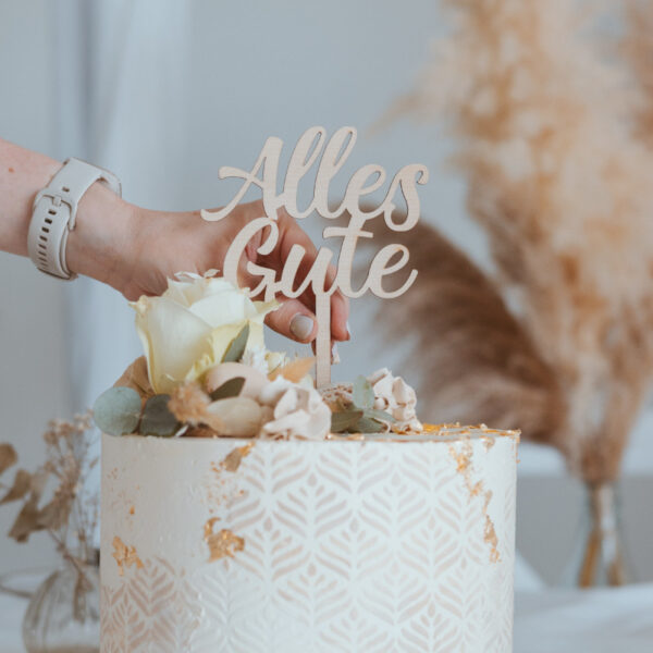 Stofftiger Caketopper Alles Gute, Hand steckt den Schriftzug in eine weiße Torte mit Rosen