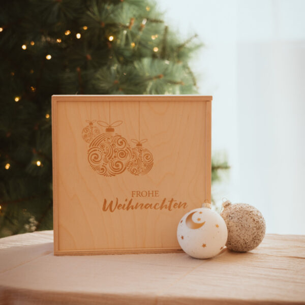 Stofftiger Holzkiste Frohe Weihnachten mit gravierten Weihnachtskugeln vor einem Christbaum